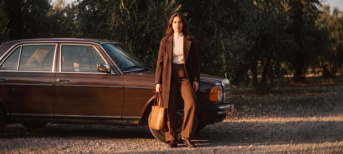 Modelo con abrigo largo marron junto a un coche clasico en un olivar al atardecer